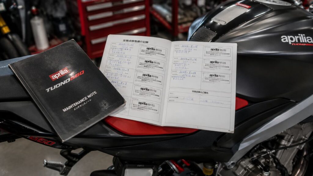 中古バイクショップに展示されたアプリリア・トゥオーノ660のシート上に広げられた、過去のメンテナンスノートや整備記録簿。